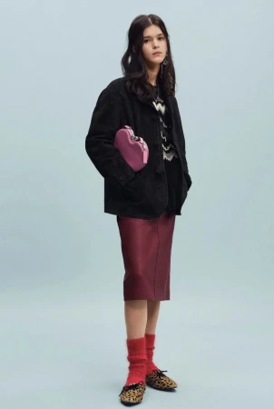 Model wearing a black jacket, burgundy midi skirt, leopard flats, and holding a pink heart bag against a light blue backdrop.