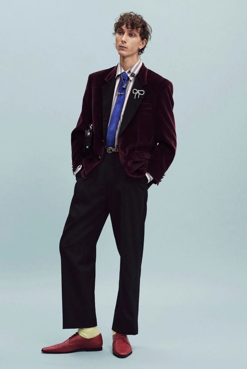 Model wearing a burgundy velvet blazer, blue tie, black trousers, and red shoes standing against a pale blue studio background.