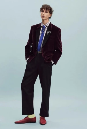 Model wearing a burgundy velvet blazer, blue tie, black trousers, and red shoes standing against a pale blue studio background.