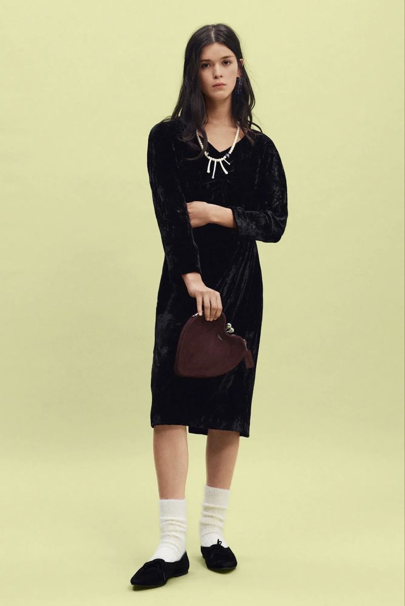 Model standing in a black velvet dress, white socks, and black flats while holding a burgundy heart bag against a pale yellow backdrop.