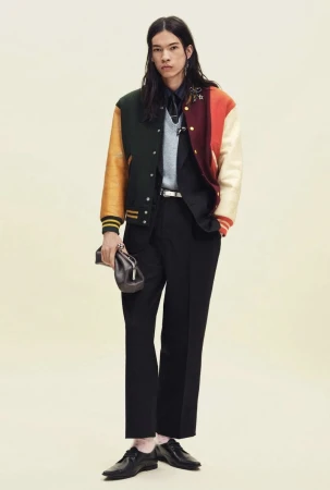 Model wearing a multi-colored varsity jacket and black tailored trousers while holding a dark clutch against a pale background.