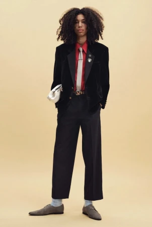 Model with voluminous curly hair wearing a black velvet blazer, red shirt, and wide-leg trousers against a warm beige studio backdrop.