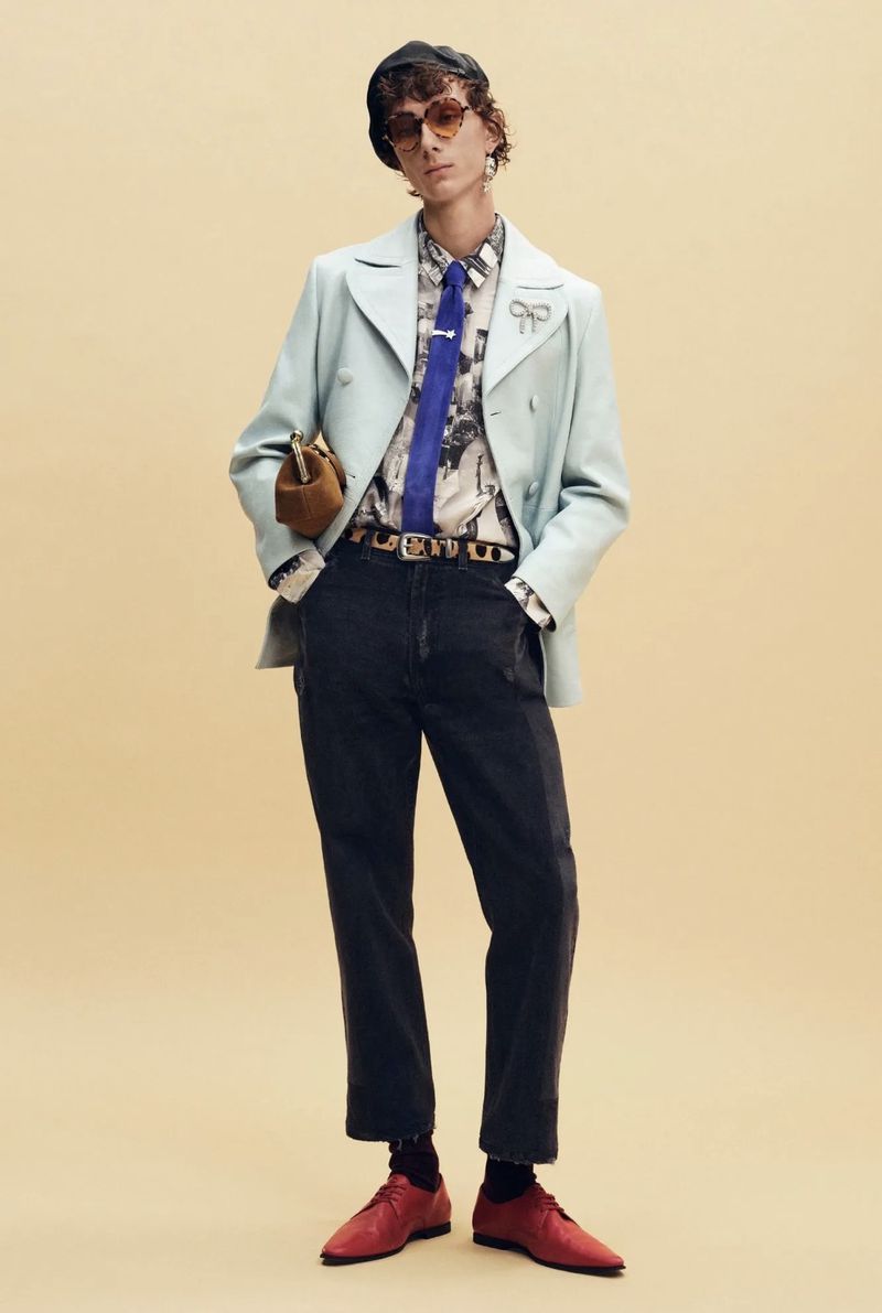 Model wearing a pale blue jacket, blue tie, dark cropped trousers, and red shoes against a beige studio backdrop for Coach.