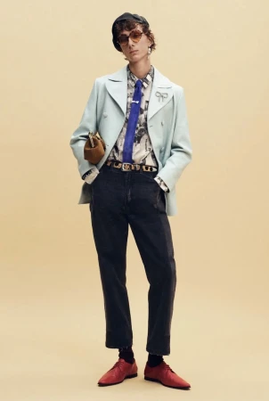 Model wearing a pale blue jacket, blue tie, dark cropped trousers, and red shoes against a beige studio backdrop for Coach.