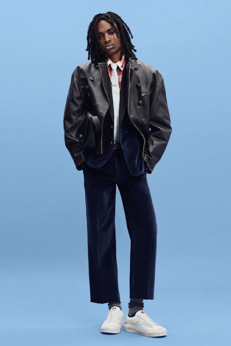 Model in an oversized black leather jacket and navy velvet trousers standing against a bright blue studio backdrop.