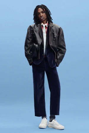 Model in an oversized black leather jacket and navy velvet trousers standing against a bright blue studio backdrop.