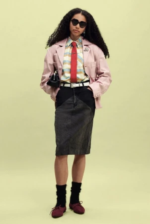 Model standing in a pink jacket, striped shirt with red tie, and dark denim skirt against a pale yellow studio backdrop.