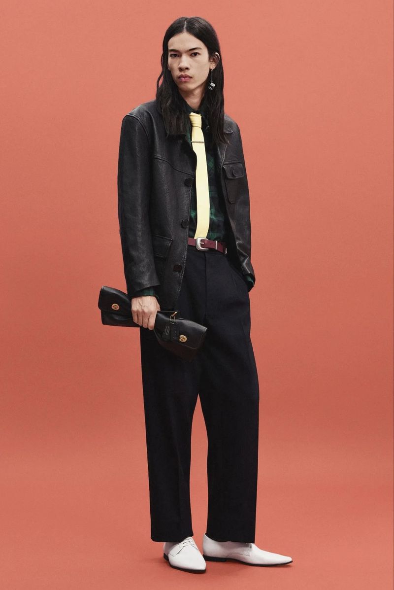 Model wearing a black leather jacket, yellow tie, wide-leg dark trousers, and white shoes against a solid coral studio backdrop.