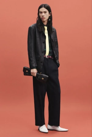 Model wearing a black leather jacket, yellow tie, wide-leg dark trousers, and white shoes against a solid coral studio backdrop.