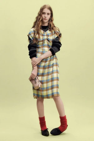 Model wearing a plaid sleeveless dress over a black knit sweater with red socks against a pale green backdrop.