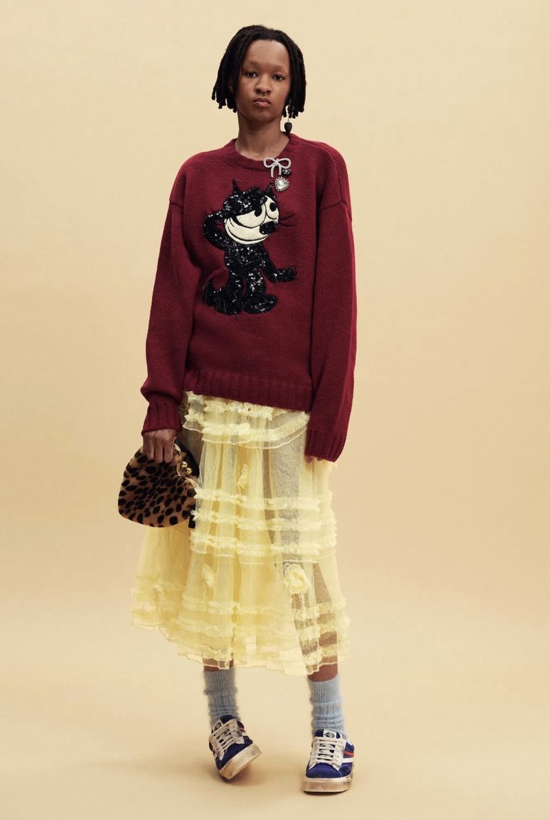Model wearing a maroon graphic cat sweater, tiered pale yellow lace skirt, sneakers, and leopard bag against a beige background.