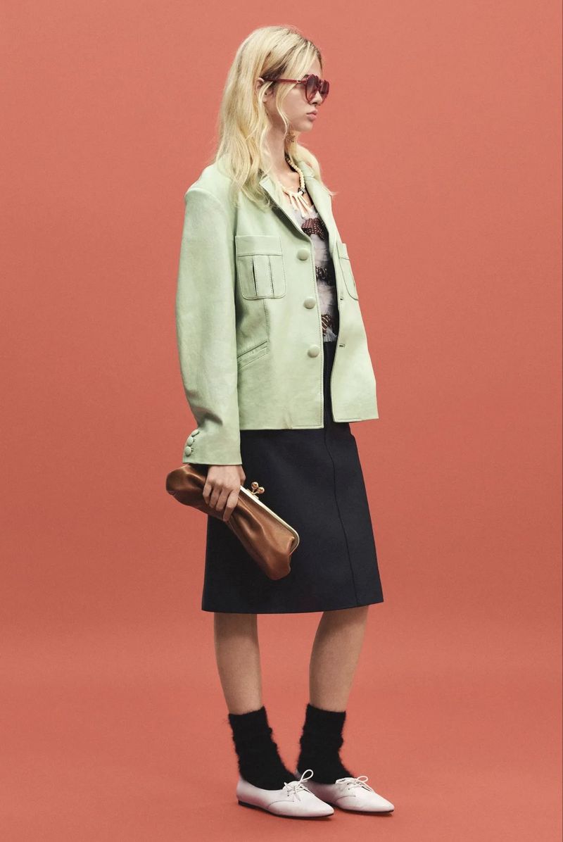 Model wearing a light green jacket, dark skirt, and holding a brown clutch against a solid red-orange seamless backdrop