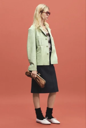 Model wearing a light green jacket, dark skirt, and holding a brown clutch against a solid red-orange seamless backdrop