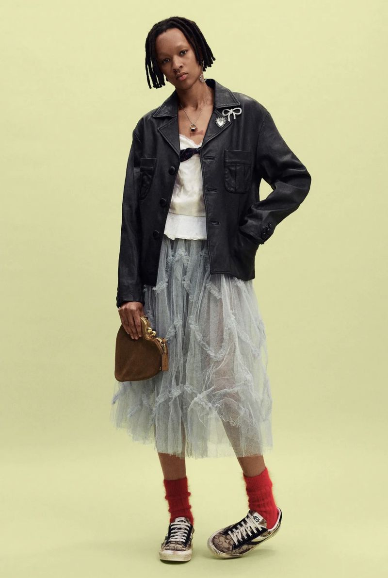 Black model wearing a black leather jacket, sheer tiered tulle skirt, red socks, and sneakers while holding a brown heart handbag.
