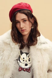 Model wearing a bright red beret and plush white faux fur coat over a graphic top against a warm studio backdrop.