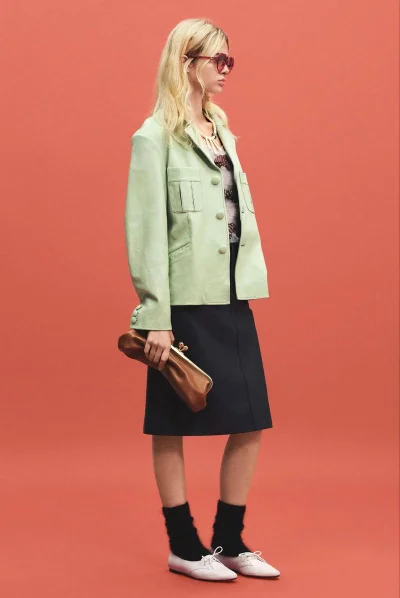 Model wearing a light green jacket, dark skirt, and holding a brown clutch against a solid red-orange seamless backdrop