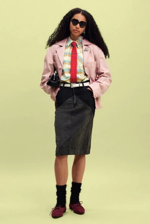 Model standing in a pink jacket, striped shirt with red tie, and dark denim skirt against a pale yellow studio backdrop.