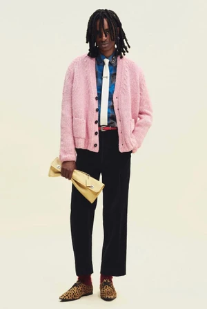 Model posing in a pink knit cardigan, black trousers, white tie, and leopard print shoes holding a yellow clutch on a light background.