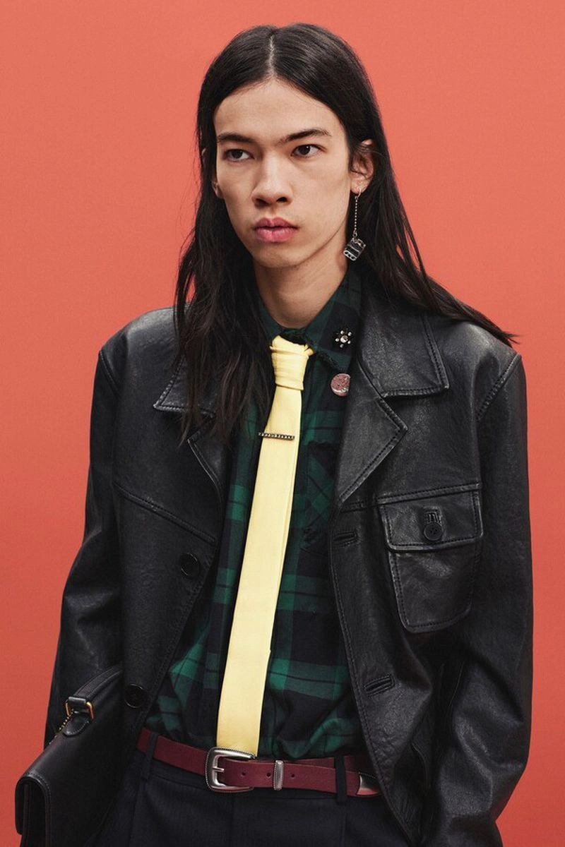 Model wearing a black leather jacket, green plaid shirt, and pale yellow tie against a terracotta background.