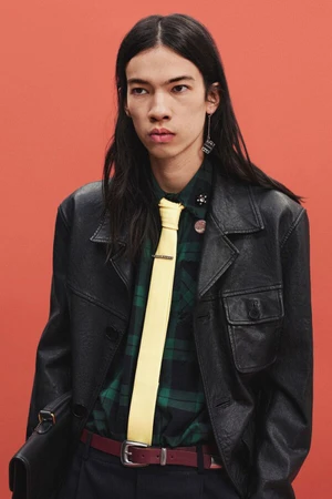Model wearing a black leather jacket, green plaid shirt, and pale yellow tie against a terracotta background.