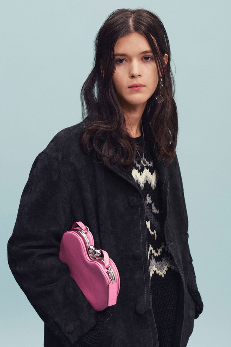 Model wearing an oversized black coat and carrying a bright pink heart-shaped purse against a pale blue seamless backdrop.