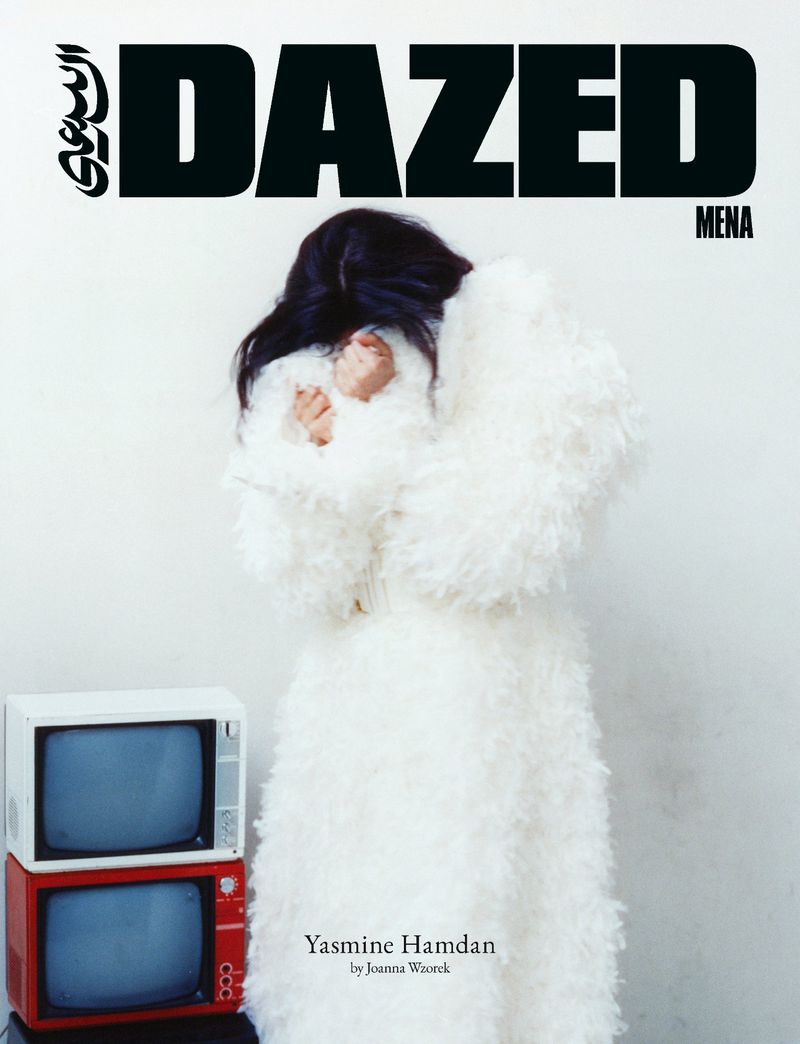 Yasmine Hamdan with hands covering her face in a white textured coat beside stacked vintage CRT televisions against a white backdrop.