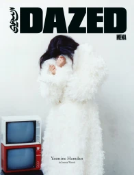 Yasmine Hamdan with hands covering her face in a white textured coat beside stacked vintage CRT televisions against a white backdrop.