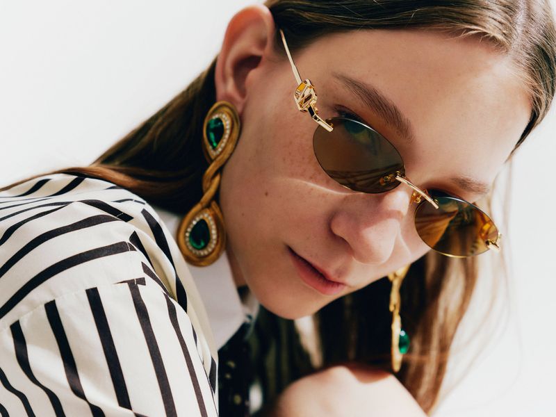 Model wearing gold oval sunglasses, large emerald teardrop earrings, and a black and white striped shirt against a white backdrop.