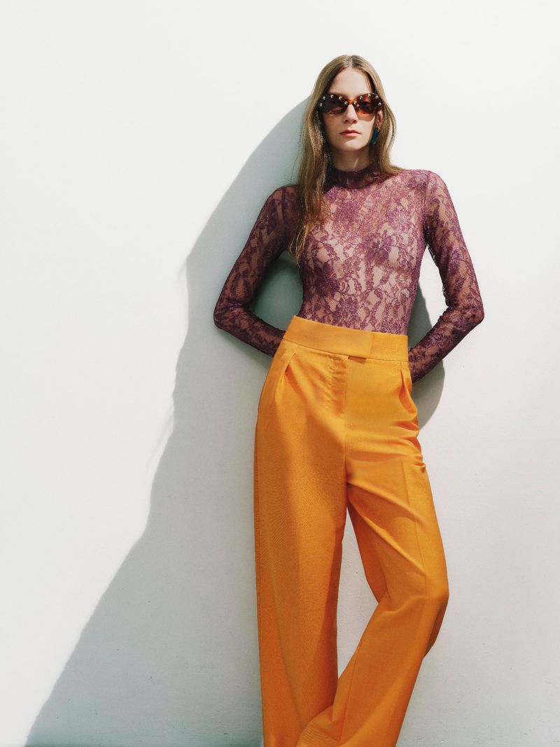 Beatrice Luzietti in a sheer purple lace top and bright orange trousers wearing sunglasses against a white backdrop with hard shadows.