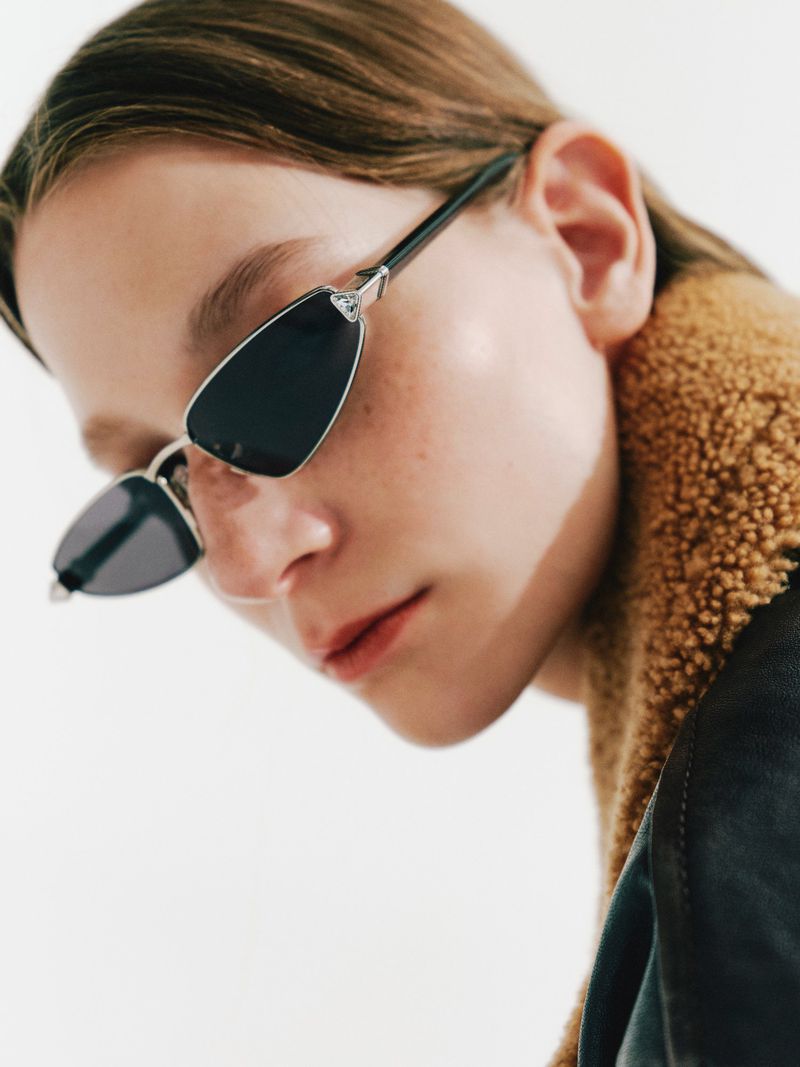 Model Beatrice Luzietti wearing slim angular black sunglasses and a shearling-collared coat against a white background.