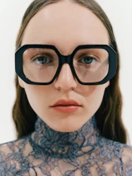 Close-up portrait of a woman wearing oversized geometric black eyeglasses and a high-neck blue lace top against a white backdrop.