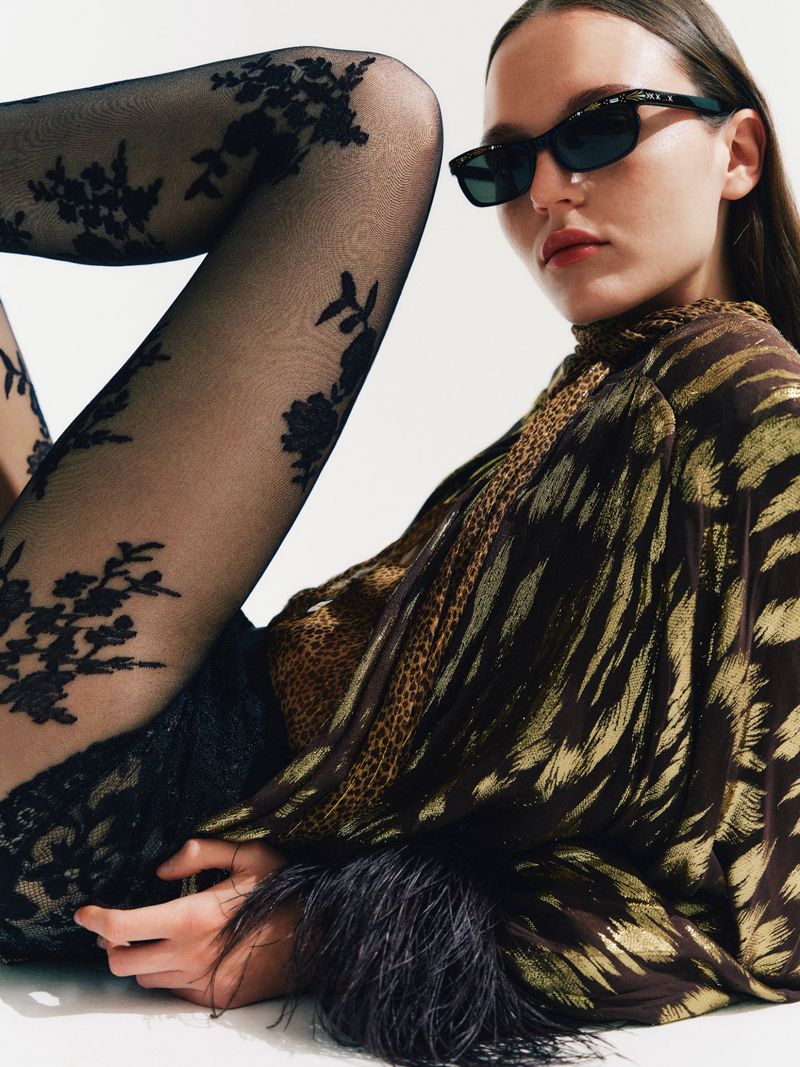 Model wearing black floral lace tights, dark sunglasses, and a metallic feather-cuffed blouse against a white background.