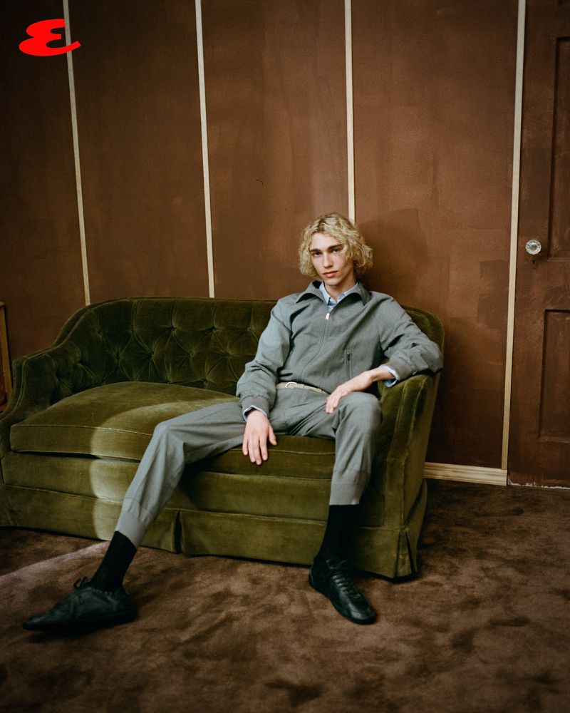 Young adult with blond curly hair in grey suit seated on green velvet couch indoors, brown walls, dark shoes and socks.