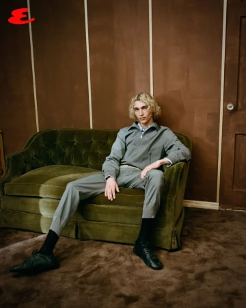 Young adult with blond curly hair in grey suit seated on green velvet couch indoors, brown walls, dark shoes and socks.