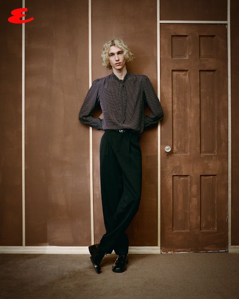 Full-length portrait of a blond male adult fashion model posing in a studio with a brown wall and a painted brown door.