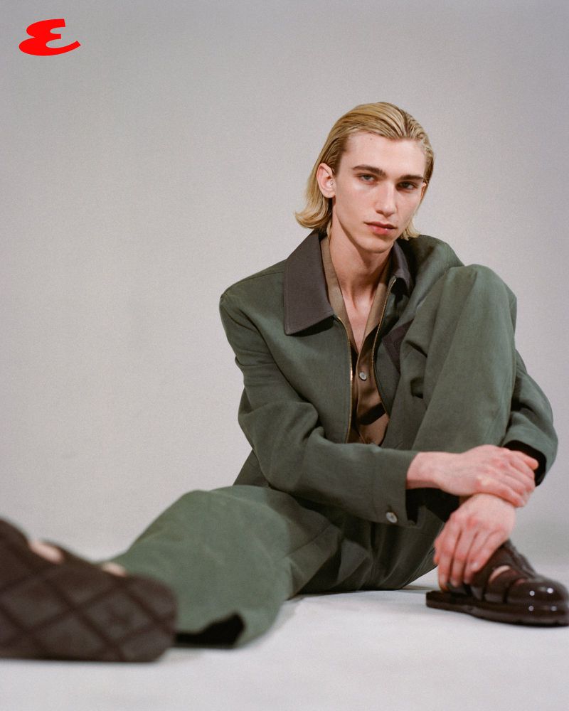 Young adult with blond hair sits against a gray studio backdrop wearing a green suit jacket and sandals for a fashion editorial shot.