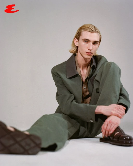 Young adult with blond hair sits against a gray studio backdrop wearing a green suit jacket and sandals for a fashion editorial shot.