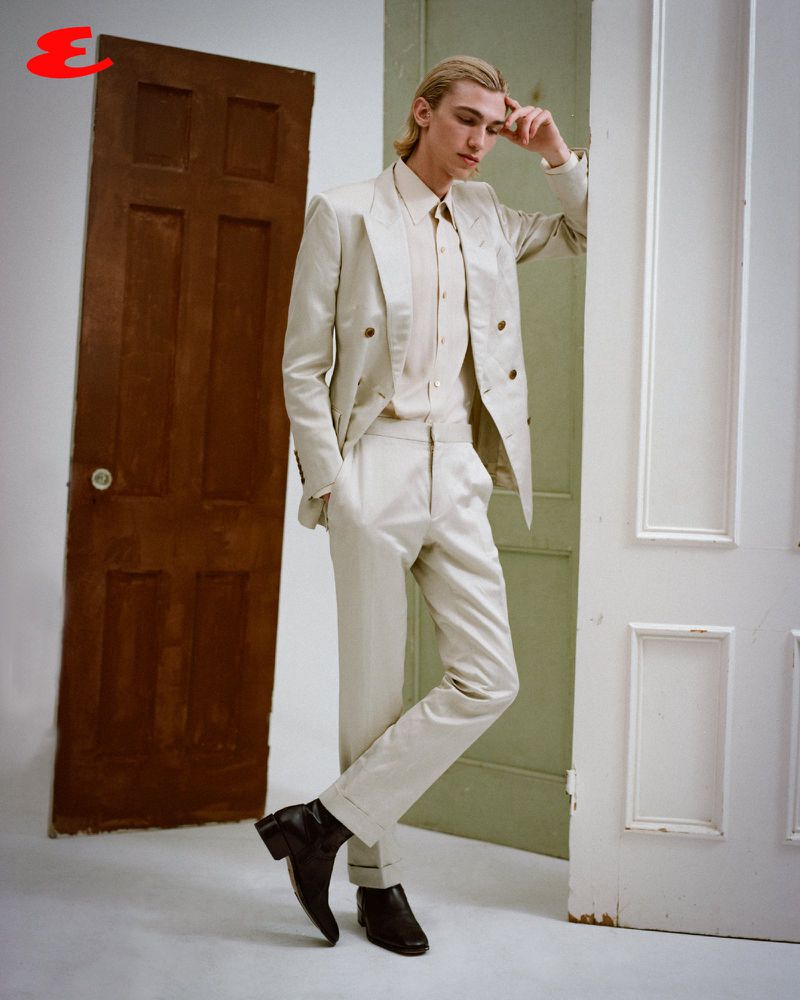 A blond male fashion model poses in a cream suit between two doorways with black Chelsea boots in a studio setting.