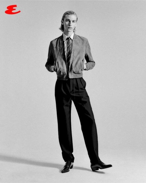 A full shot of a tall, slim male model in a grey jacket, white shirt, black tie and black trousers, posing in a photo studio for a fashion editorial.