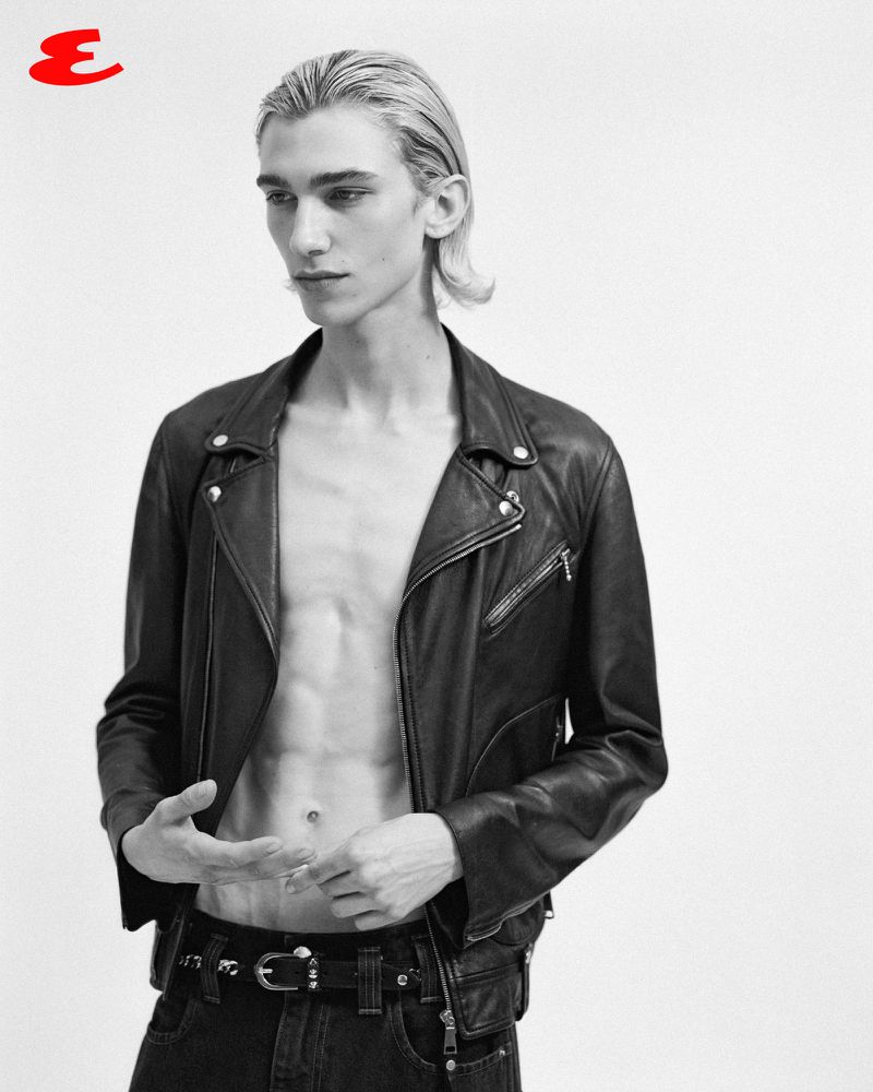 Black and white studio portrait of a fair-skinned young adult male model in an open black leather jacket and dark jeans. Esquire logo.