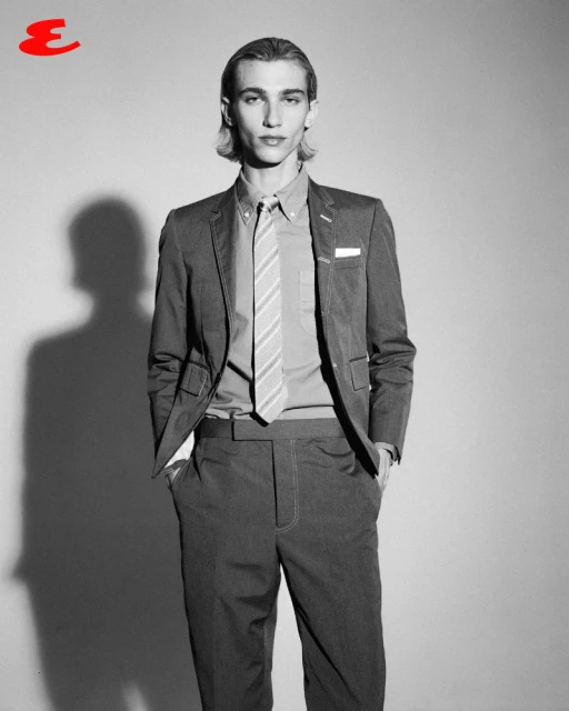 Black and white portrait of a male model in a suit and tie standing in front of a light gray wall in a photo studio setting.