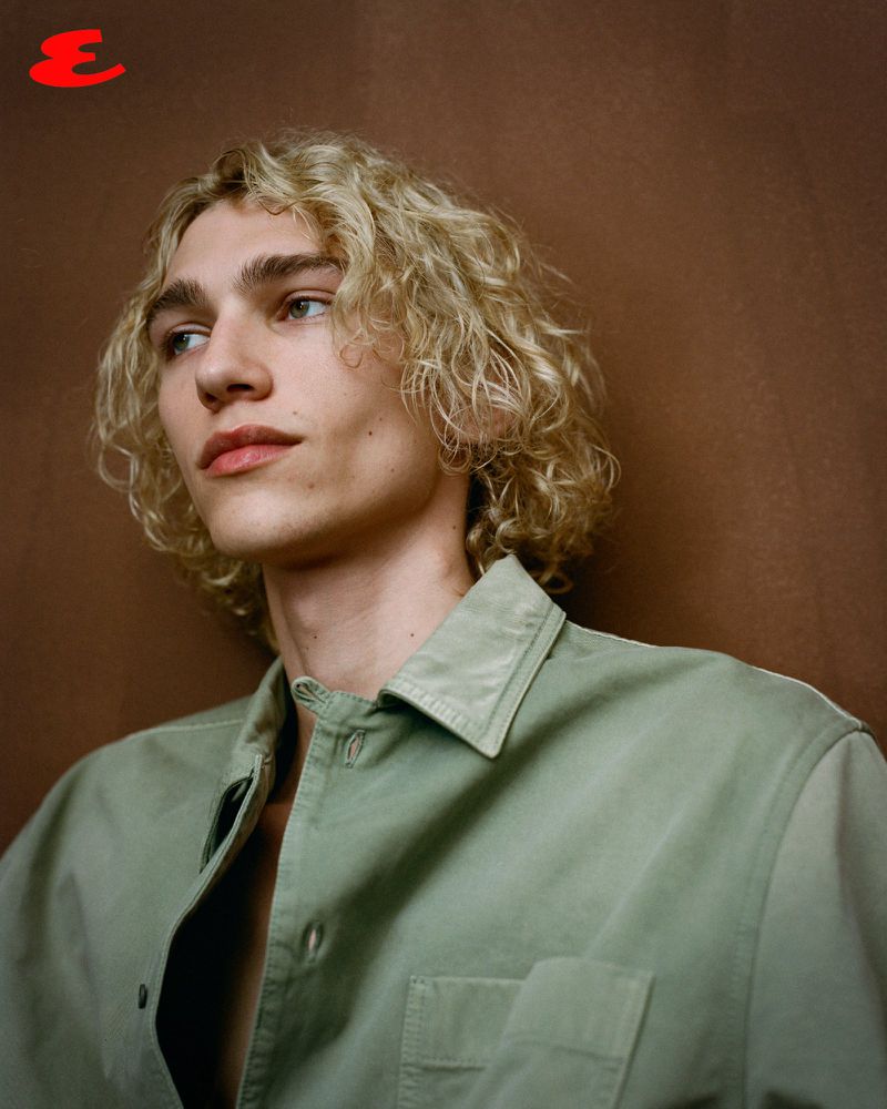 Portrait of a young blond male model in a casual sage-green button-down shirt against a brown studio backdrop with soft, diffused lighting.