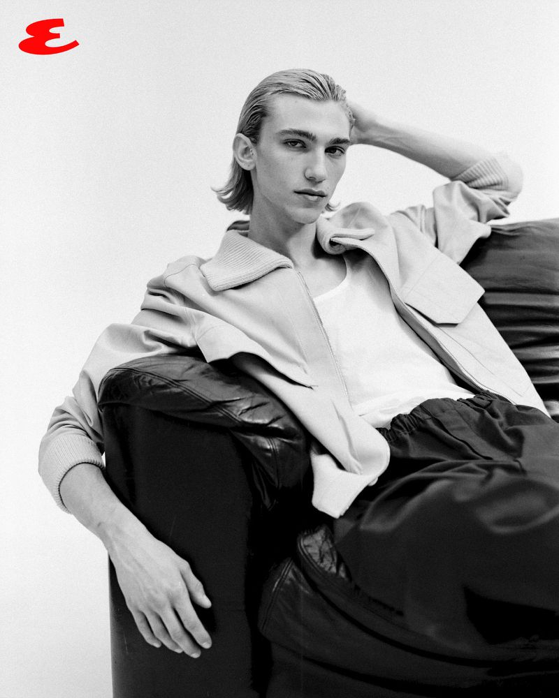 Monochrome portrait of a young man with blond hair lounging on a black leather sofa, wearing a jacket and tank top in a photo studio.
