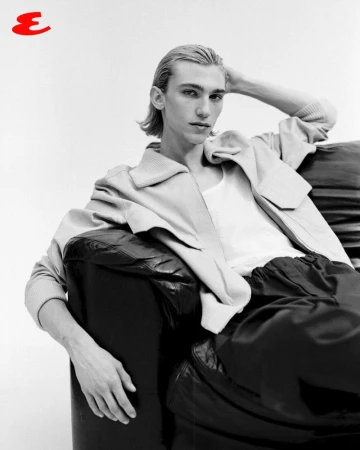 Monochrome portrait of a young man with blond hair lounging on a black leather sofa, wearing a jacket and tank top in a photo studio.