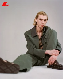 Model Noah Hanes wearing an olive green jacket, matching trousers, and brown leather sandals seated against a studio backdrop.