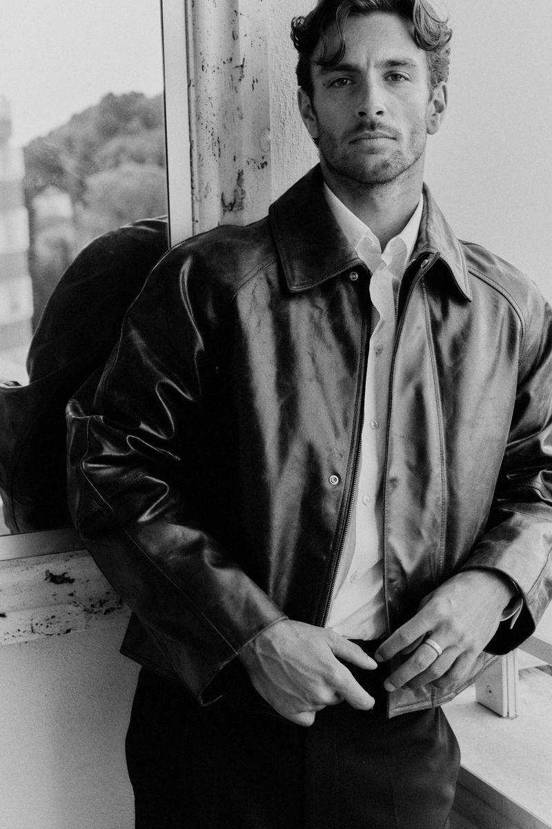 Lorenzo Musetti in a black leather jacket and white collared shirt posing beside a window in a black and white portrait.