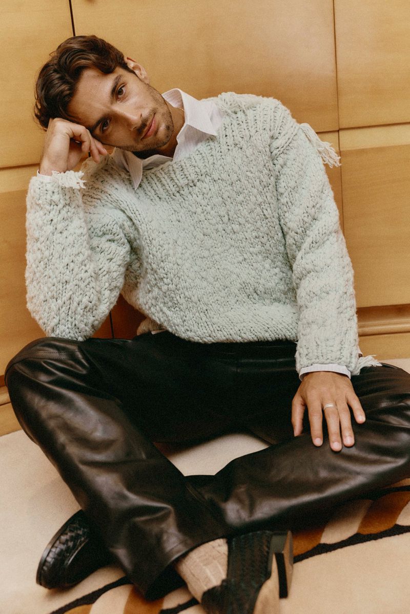 Lorenzo Musetti seated on the floor wearing a mint green textured knit sweater and black leather trousers against wood panelling.