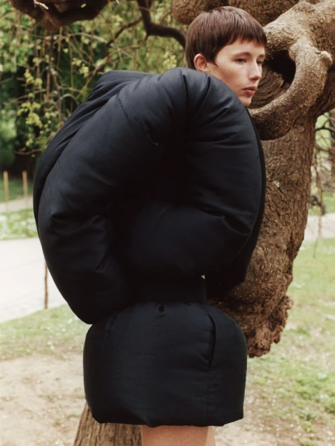 Fashion portrait of a person with short hair, wearing a black padded jacket and skirt, posing next to a tree in an outdoor setting.