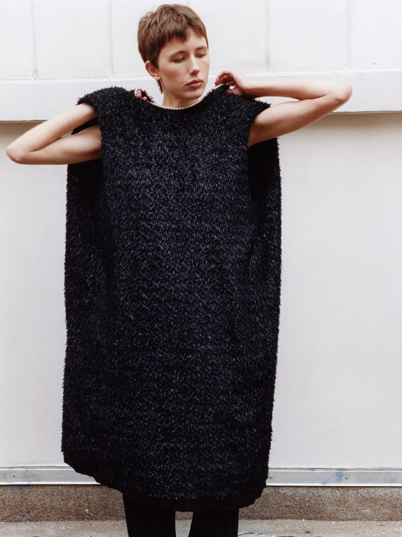 A young adult female in a black textured poncho-style garment poses against a white wall indoors, looking slightly downward with hands raised.