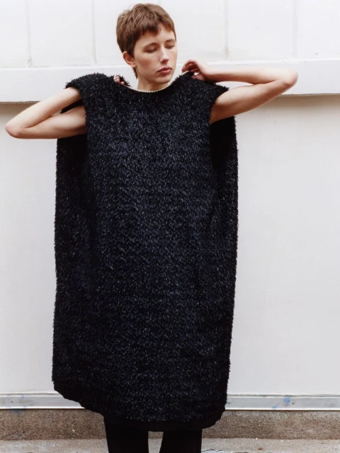A young adult female in a black textured poncho-style garment poses against a white wall indoors, looking slightly downward with hands raised.